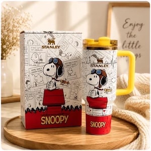 Könnte beinhalten: Ein weiß-roter Stanley-Becher mit gelbem Henkel und Deckel, verziert mit einer Grafik von Snoopy mit Fliegerbrille auf einer roten Hundehütte. Die passende Verpackung zeigt dieselbe Snoopy-Figur sowie den Schriftzug Stanley und Snoopy.