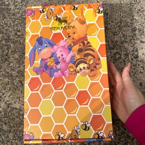 May include: A rectangular product box featuring a yellow and orange honeycomb pattern with illustrations of Winnie the Pooh, Piglet, Eeyore, and Tigger. The word STANLEY is printed in gold near the top, surrounded by small bees and honey pots.