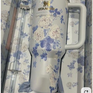 May include: A light blue Stanley tumbler with a white floral pattern and the text "LOVESHACKFANCY" printed on the front. The tumbler features a large side handle, a metallic gold logo, and a clear plastic straw, all presented in a matching floral box.