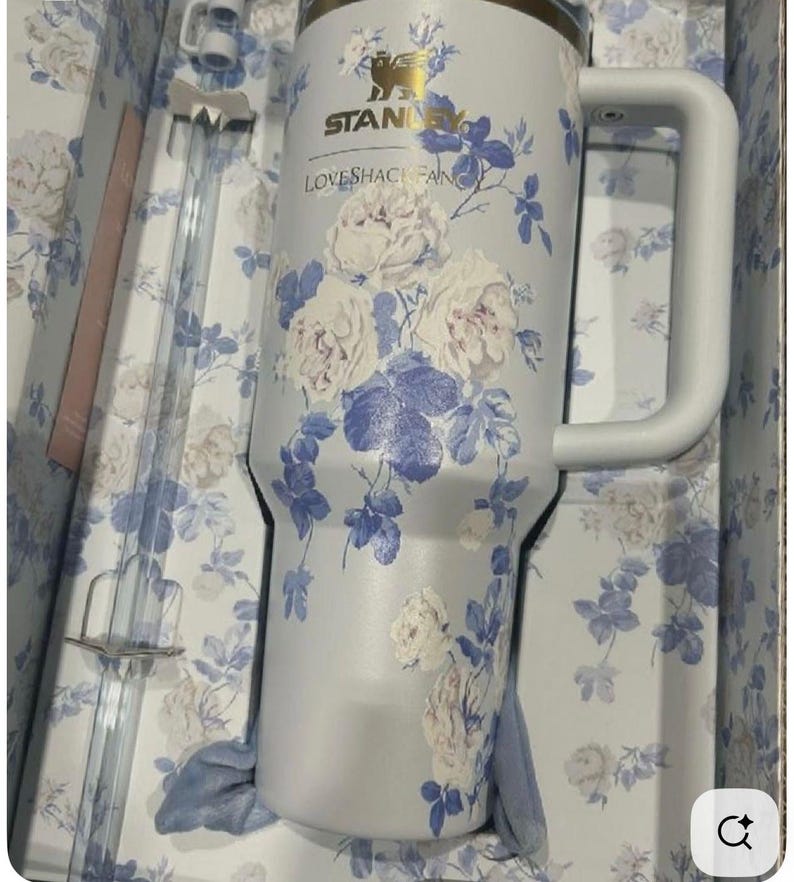 May include: A light blue Stanley tumbler with a white floral pattern and the text "LOVESHACKFANCY" printed on the front. The tumbler features a large side handle, a metallic gold logo, and a clear plastic straw, all presented in a matching floral box.