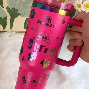 May include: A bright pink Stanley tumbler featuring a handle, a translucent pink lid, and a repeating pattern of the character Stitch, hibiscus flowers, and stars. The word Stitch is printed in large letters, and the Stanley logo is visible.