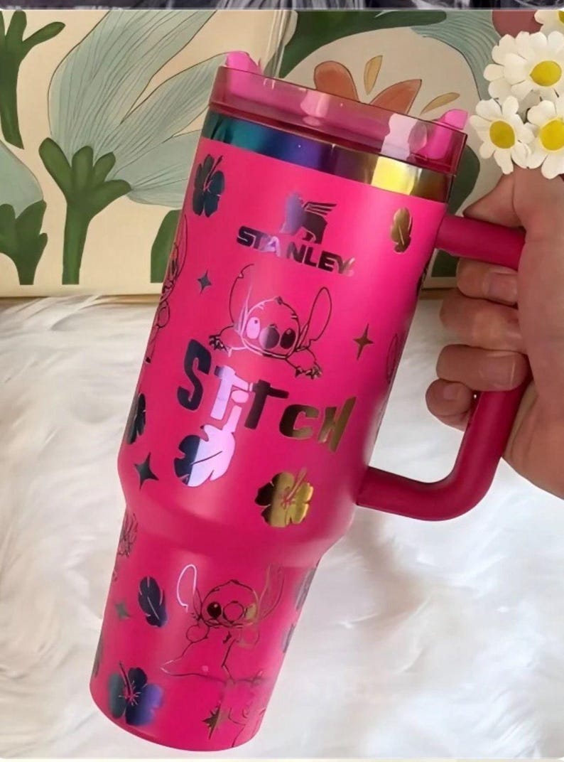 May include: A bright pink Stanley tumbler featuring a handle, a translucent pink lid, and a repeating pattern of the character Stitch, hibiscus flowers, and stars. The word Stitch is printed in large letters, and the Stanley logo is visible.