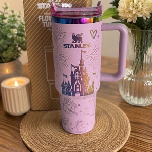 May include: A light purple Stanley tumbler featuring a metallic rainbow-colored castle graphic, fireworks, and various character illustrations. The cup includes a matching purple handle, a clear lid, and the Stanley logo printed on the front.
