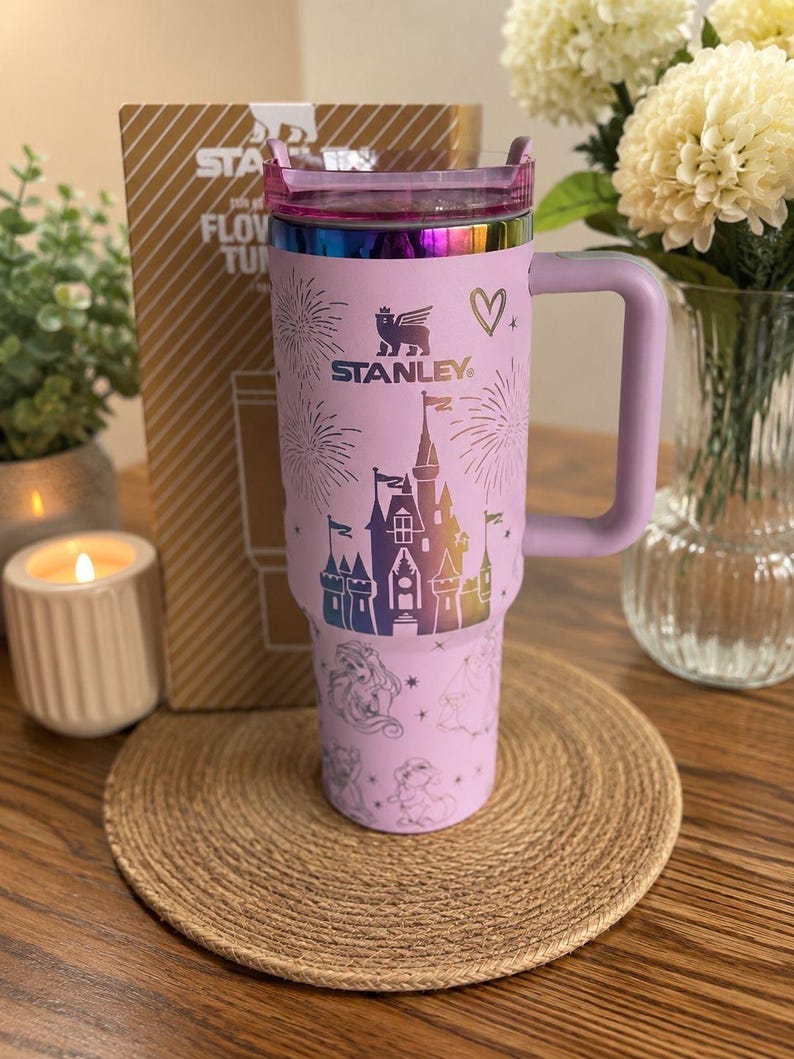 May include: A light purple Stanley tumbler featuring a metallic rainbow-colored castle graphic, fireworks, and various character illustrations. The cup includes a matching purple handle, a clear lid, and the Stanley logo printed on the front.