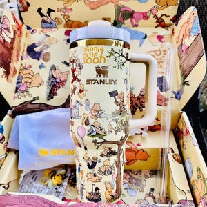 May include: A Stanley Winnie the Pooh themed tumbler with a white handle and lid, and a gold rim. The tumbler is covered in illustrations of Winnie the Pooh characters. The tumbler is in a matching box with a straw and a blue cloth.