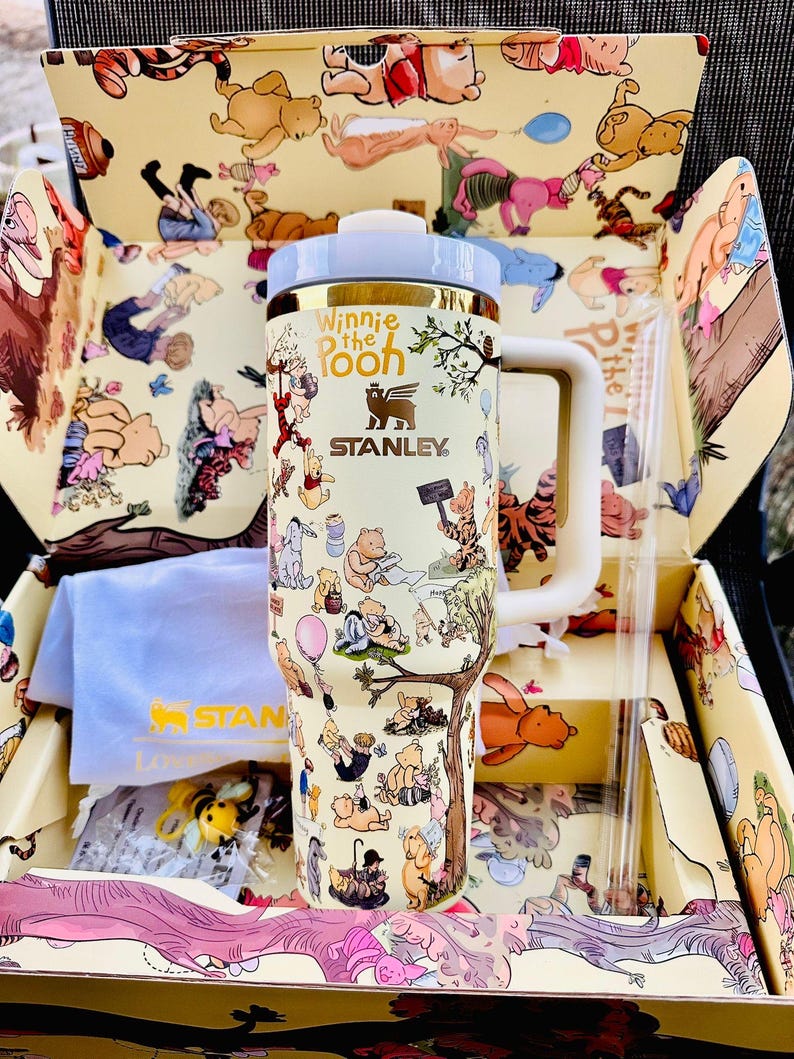 May include: A Stanley Winnie the Pooh themed tumbler with a white handle and lid, and a gold rim. The tumbler is covered in illustrations of Winnie the Pooh characters. The tumbler is in a matching box with a straw and a blue cloth.