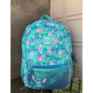 Puede incluir: Una mochila de color verde azulado con un patr&oacute;n repetitivo del personaje Stitch de Disney, intercalado con flores de hibisco rosas y hojas tropicales verdes. Un bolsillo frontal azul met&aacute;lico muestra un gr&aacute;fico de Stitch y una etiqueta Disney Smiggle.