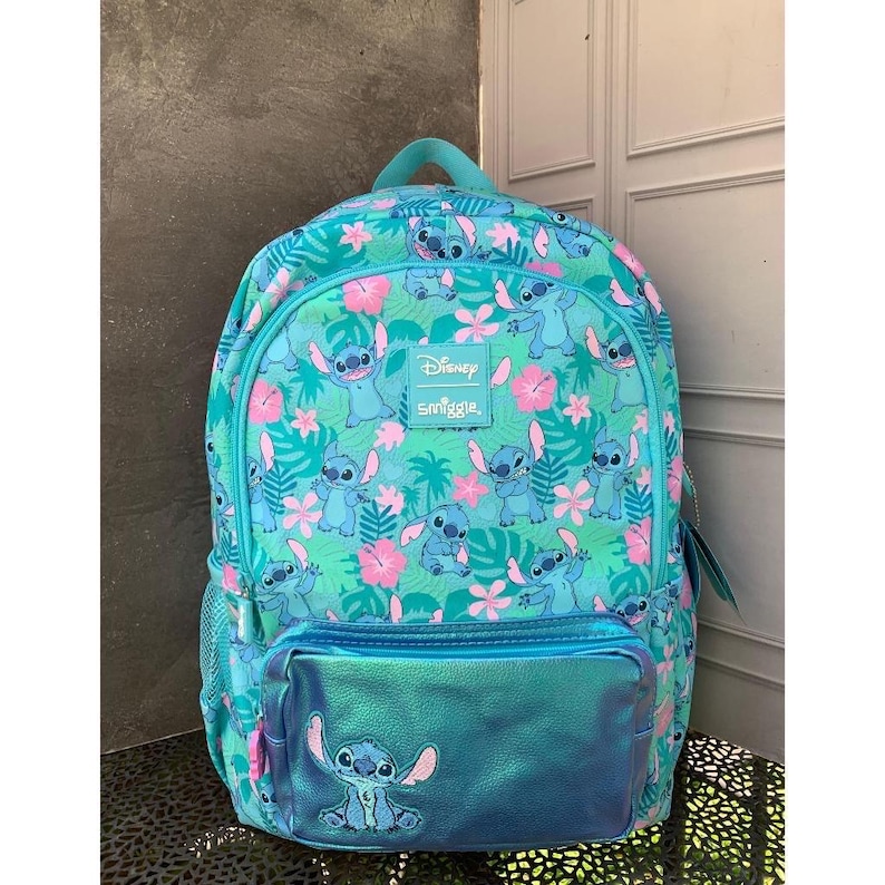 Puede incluir: Una mochila de color verde azulado con un patr&oacute;n repetitivo del personaje Stitch de Disney, intercalado con flores de hibisco rosas y hojas tropicales verdes. Un bolsillo frontal azul met&aacute;lico muestra un gr&aacute;fico de Stitch y una etiqueta Disney Smiggle.