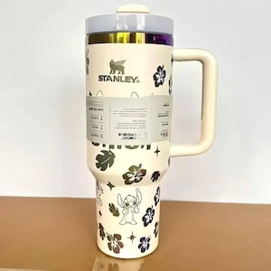 May include: A cream-colored Stanley tumbler with a large handle and a clear lid. The exterior features a repeating pattern of dark hibiscus flowers, small stars, and the character Stitch. A paper product label wraps around the center of the cup.
