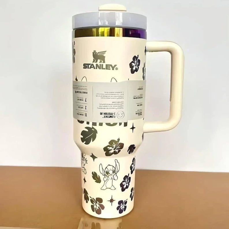 May include: A cream-colored Stanley tumbler with a large handle and a clear lid. The exterior features a repeating pattern of dark hibiscus flowers, small stars, and the character Stitch. A paper product label wraps around the center of the cup.