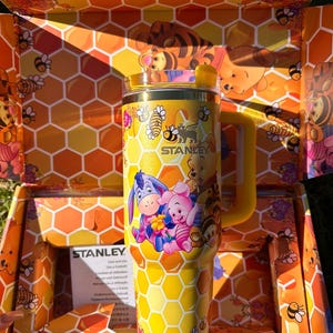 May include: A yellow Stanley tumbler featuring a honeycomb pattern and illustrations of Winnie the Pooh, Piglet, Eeyore, and Tigger. The cup has a yellow handle, a clear lid with a yellow straw opening, and is presented in a matching honeycomb box.