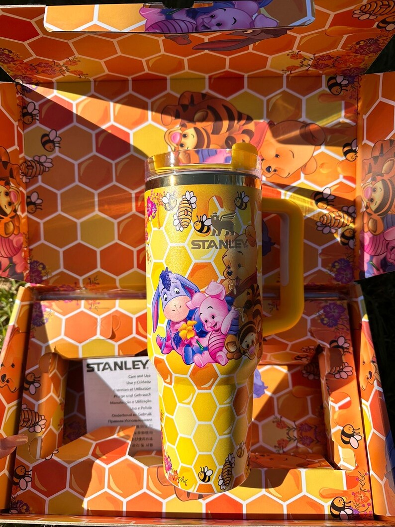 May include: A yellow Stanley tumbler featuring a honeycomb pattern and illustrations of Winnie the Pooh, Piglet, Eeyore, and Tigger. The cup has a yellow handle, a clear lid with a yellow straw opening, and is presented in a matching honeycomb box.
