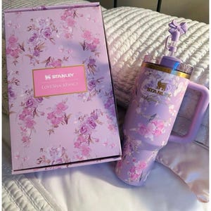 May include: A lavender Stanley tumbler with a floral pattern, a handle, and a straw topped with a bow, accompanied by a matching rectangular box. Both items feature the Stanley and LoveShackFancy logos printed in gold on a light purple floral background.