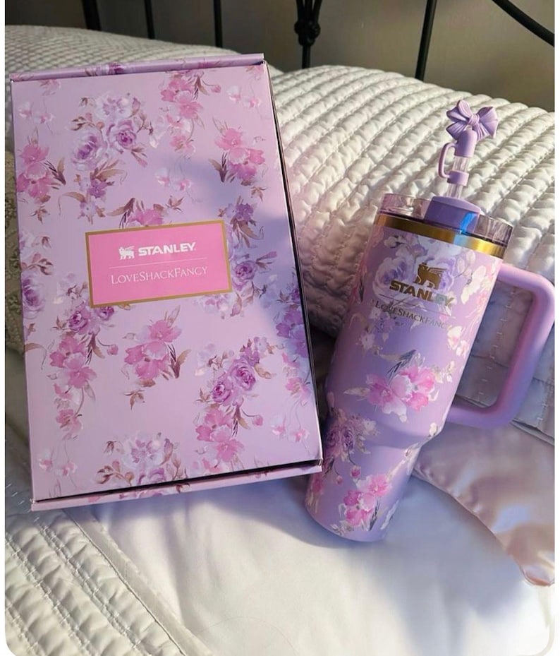 May include: A lavender Stanley tumbler with a floral pattern, a handle, and a straw topped with a bow, accompanied by a matching rectangular box. Both items feature the Stanley and LoveShackFancy logos printed in gold on a light purple floral background.