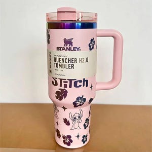 May include: A light pink 40 oz Stanley Flowstate Quencher H2.0 tumbler with a handle, a lid, and a rainbow metallic rim. The body features dark purple hibiscus flower patterns, star icons, and the character Stitch with the text STITCH.