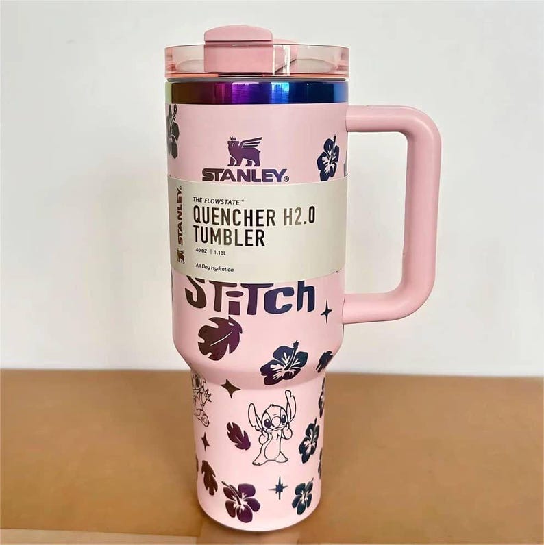 May include: A light pink 40 oz Stanley Flowstate Quencher H2.0 tumbler with a handle, a lid, and a rainbow metallic rim. The body features dark purple hibiscus flower patterns, star icons, and the character Stitch with the text STITCH.