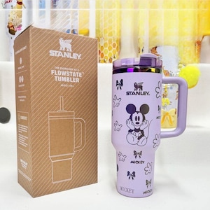 May include: A light purple Stanley Quencher H2.0 FlowState tumbler with a 40 oz capacity, featuring a Mickey Mouse graphic, bows, and glove prints. The tumbler includes a handle, a purple lid, and comes with its original brown cardboard box.