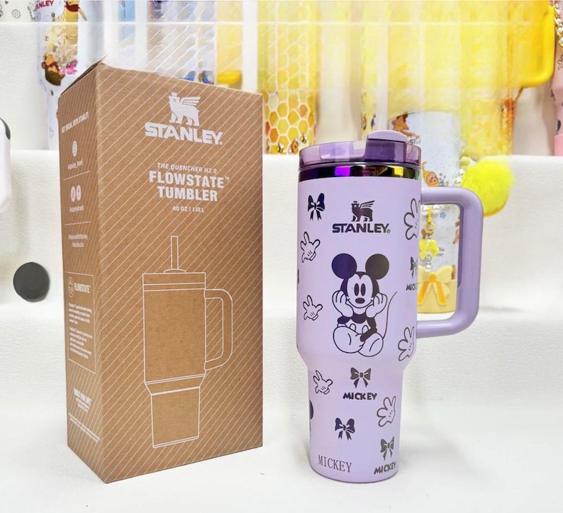 May include: A light purple Stanley Quencher H2.0 FlowState tumbler with a 40 oz capacity, featuring a Mickey Mouse graphic, bows, and glove prints. The tumbler includes a handle, a purple lid, and comes with its original brown cardboard box.