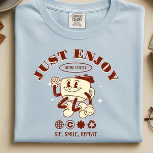 Puede incluir: Una camiseta azul claro con un gráfico de una taza de café de dibujos animados con brazos y piernas. El texto dice JUST ENJOY SOME COFFEE y SIP, SMILE, REPEAT. Debajo de la taza hay iconos de un globo terráqueo, copyright, una flor y reciclaje.