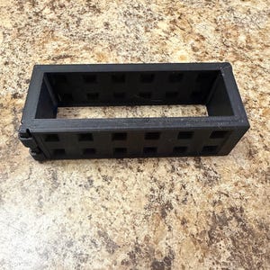 May include: A rectangular black plastic frame featuring a grid-like pattern of square cutouts along its four sides. The structure is open at the top and bottom, with a visible hinge mechanism on the left side connecting the frame segments together.