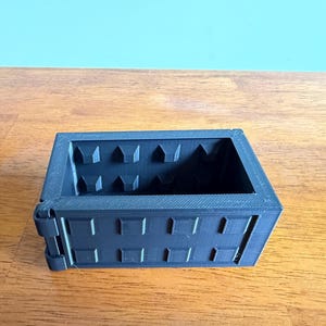 Puede incluir: Un contenedor de almacenamiento rectangular negro con un centro hueco y un exterior texturizado que presenta una rejilla de tacos cuadrados elevados. El contenedor tiene un panel lateral con bisagras y un marco rígido con acabado 3D visible.