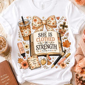 May include: A white t-shirt featuring a graphic print of an open book with the text "SHE IS CLOTHED IN STRENGTH Proverbs 31:25". The design includes a floral bow, an iced coffee cup, a highlighter, a small notebook, a pen, and a cross.
