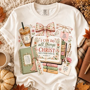 May include: A cream-colored t-shirt featuring a graphic print of an open Bible with the text I CAN DO all things THROUGH CHRIST WHO STRENGTHENS ME, a floral bow, a coffee cup, a notebook, eyeglasses, highlighters, and inspirational notes.