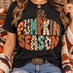 May include: A black short-sleeved t-shirt featuring the text PUMPKIN SEASON in large, block-style letters. Each letter is filled with various patterns including stars, cow print, turquoise, paisley, and checkered designs, topped with a cowboy hat.