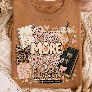 May include: A tan t-shirt featuring a graphic print with the text "Pray MORE Worry LESS". The design includes a prayer journal, a leopard print bow, an iced coffee, a Holy Bible, a pen, and a note with "Be still and know that I am God".