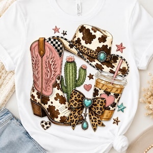 May include: A white t-shirt featuring a graphic print of a pink and brown cow print cowboy boot, a cow print cowboy hat with a leopard print band, a green cactus, a leopard print bow with a turquoise heart charm, and an iced coffee cup.
