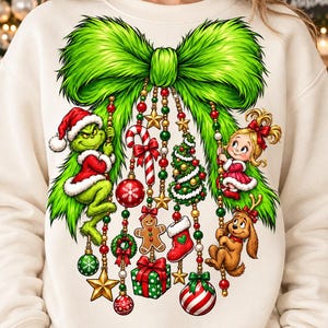May include: A cream-colored sweatshirt featuring a large, vibrant green fur-textured bow graphic. Hanging from the bow are strings of beads with ornaments including a green character, a blonde girl, a brown dog, a candy cane, a gingerbread man, and gifts.