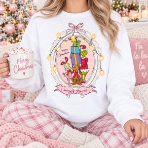 Op de afbeelding: Een wit sweatshirt met ronde hals en een afbeelding van de Grinch, Cindy Lou Who en Max die cadeaus vasthouden in een roze ovaal frame versierd met kerstverlichting en een strik. Een witte mok met de tekst Merry Christmas is ook afgebeeld.