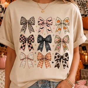 May include: A beige t-shirt featuring a grid of nine decorative bows with various patterns, including leopard print, bats, pumpkins, pink and black checkers, spider webs, floral designs, ghosts, orange plaid, and black and white cow print.