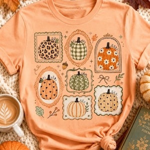 May include: A peach-coloured short-sleeve t-shirt featuring a graphic print of seven pumpkins, each enclosed in a unique decorative frame. The pumpkins display various patterns including leopard print, green gingham, white daisies, black hearts, and polka dots.