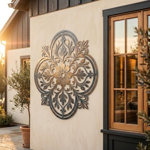Könnte beinhalten: Ein großes, ovales Wandkunstobjekt aus Metall mit einem komplexen, symmetrischen Blumen- und Blattmuster. Das Design weist ein Farbverlauf-Finish auf, das von einem dunklen, patinierten Bronze- oder Anthrazitrand zu einer warmen Goldmitte übergeht.