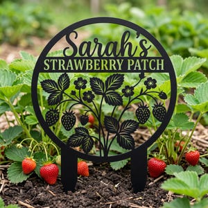 May include: A black circular garden sign with two stakes featuring a cutout design of strawberry plants and the text Sarah's STRAWBERRY PATCH. The sign is constructed with a solid frame and detailed silhouettes of leaves and berries.