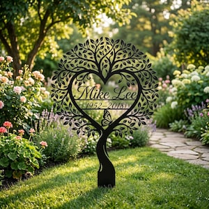 Puede incluir: Un estaca de jardín de metal negro con un diseño de árbol circular y un centro en forma de corazón. El texto "Mike Lee" y "1952-2018" está grabado en el medio, acompañado por una pequeña silueta de mariposa en el tronco inferior.