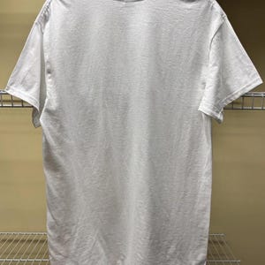 May include: A plain white short-sleeved t-shirt shown from the back. The garment features a standard crew neckline, short sleeves, and a straight hemline. The fabric appears to be a solid, opaque white material with no visible patterns, prints, or logos.
