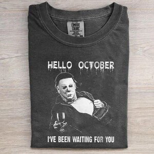 May include: A charcoal gray t-shirt featuring a white graphic of a masked character holding a wine glass. The text HELLO OCTOBER is printed above the graphic, and the text I'VE BEEN WAITING FOR YOU is printed below. The shirt has a crew neckline.