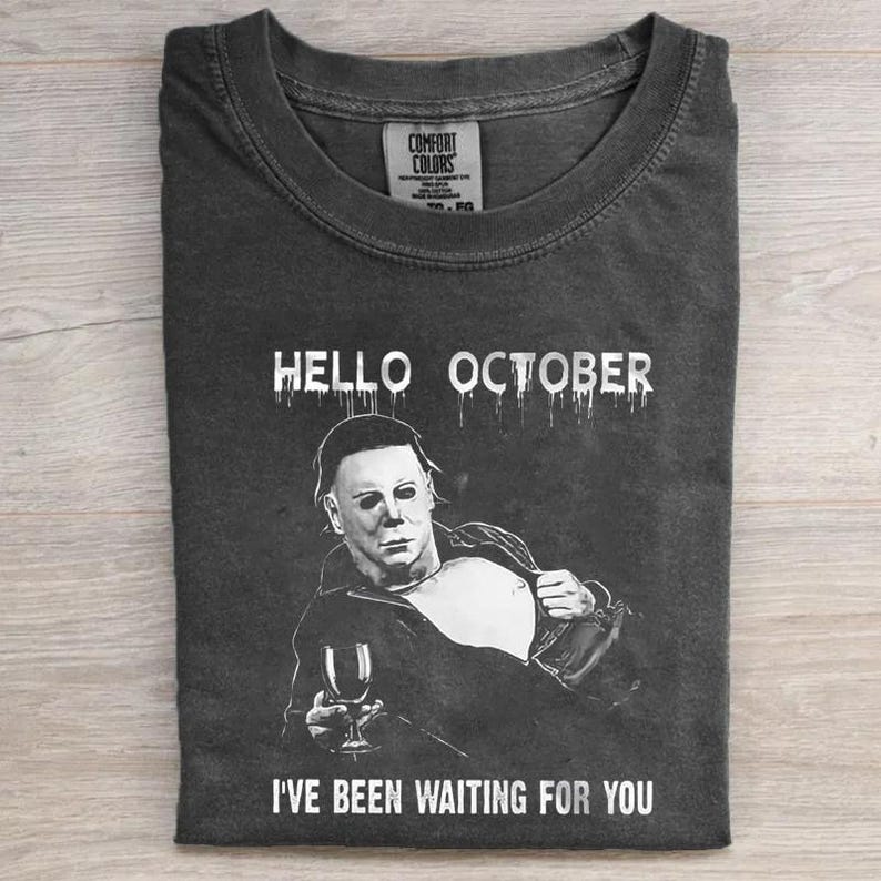 May include: A charcoal gray t-shirt featuring a white graphic of a masked character holding a wine glass. The text HELLO OCTOBER is printed above the graphic, and the text I'VE BEEN WAITING FOR YOU is printed below. The shirt has a crew neckline.