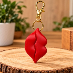 Puede incluir: Un llavero de color rojo brillante con forma de labios. El dije está unido a una argolla de metal dorado y a un cierre de mosquetón dorado. Los labios tienen un acabado mate suave y una línea vertical distintiva en el centro.