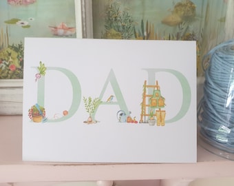 May include: A white card with the word "DAD" written in green letters. Each letter is decorated with a small illustration of gardening items, such as a watering can, a ladder, and a bird.