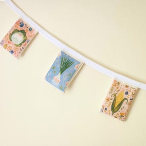 May include: A decorative banner featuring three rectangular fabric panels with illustrations of vegetables. The panels are attached to a white ribbon. The vegetables include a cauliflower, an onion, and an ear of corn, each set against a floral background.
