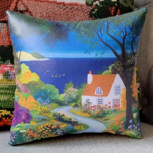 May include: Square decorative pillow featuring a scenic landscape with a white cottage, orange roof, and a winding road leading to the sea. The pillow has a vibrant color palette with blues, greens, yellows, and purples, creating a picturesque scene.