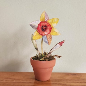 May include: A paper daffodil in a terracotta pot. The flower features yellow petals with patchwork designs, a red centre, and green leaves. The pot rests on a wooden surface, creating a decorative piece for the home.