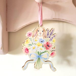 May include: Wooden Easter ornament featuring a watercolor illustration of a bunny surrounded by a bouquet of flowers. The flowers include daisies, poppies, and lavender, tied with a blue ribbon. A pink ribbon is attached for hanging.