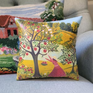 May include: Square decorative pillow with a colorful illustration of a woman picking apples from a tree in an orchard. The pillow has a light blue border and is placed on a gray sofa.