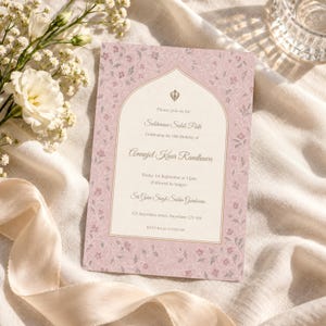 May include: A rectangular invitation card with a dusty pink border featuring a delicate floral pattern. The center is cream-colored with an arched frame, displaying text for a Sukhmani Sahib Path celebration for Amanjot Kaur Randhawa.