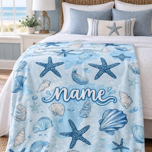 May include: A light blue throw blanket featuring a repeating pattern of dark blue starfish and various white seashells. The centre of the blanket displays the word name in a stylised, cursive blue font, set against a soft watercolour background.