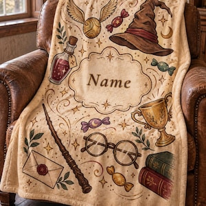 May include: A beige fleece blanket featuring a central decorative frame with the text Name, surrounded by illustrations of a winged golden ball, a brown pointed hat, a potion bottle, a wand, round glasses, a gold trophy, books, and sweets.